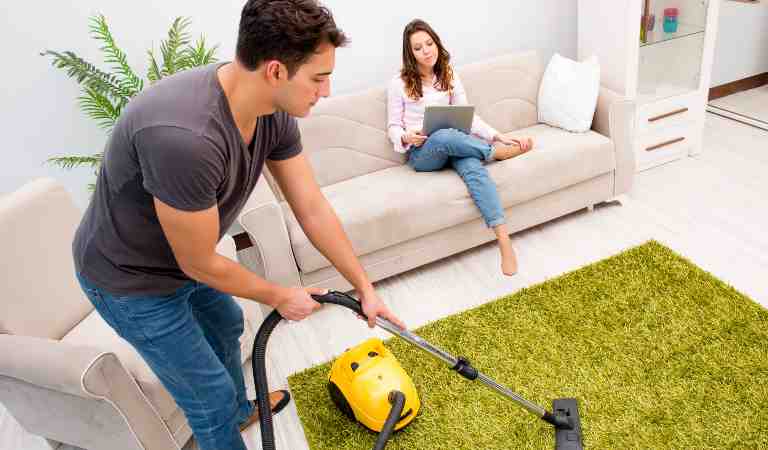 A woman is sitting on sofa and a man is vacuuming carpet inside a living room.
