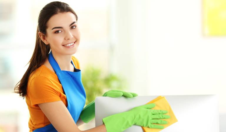 Woman in orange-blue uniform and green glove cleaning computer inside a office with a yellow cloth.