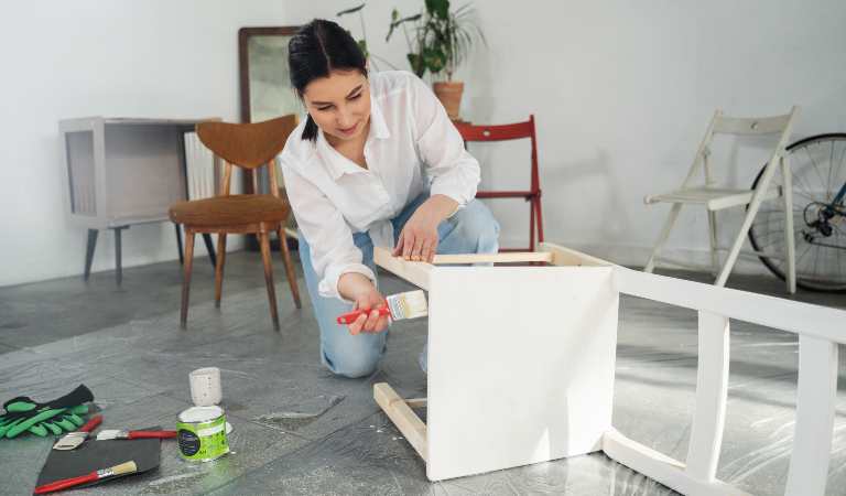 Woman in white dress painting her old furniture using a white paint.