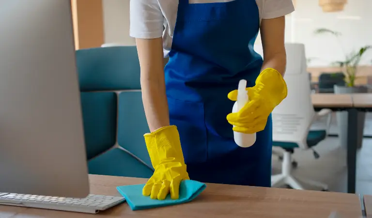 Woman in white-blue uniform and yellow gloves cleaning a table in a office.