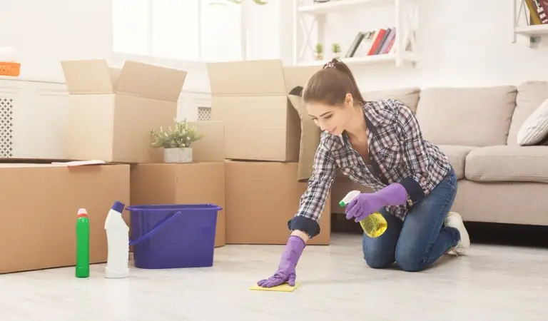 Woman in check shirt cleaning floor and some boxes, one green bottle, one white bottle and a purple basket on the floor.