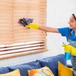 Smiling professional cleaner in blue uniform wipes window blinds in a modern living room