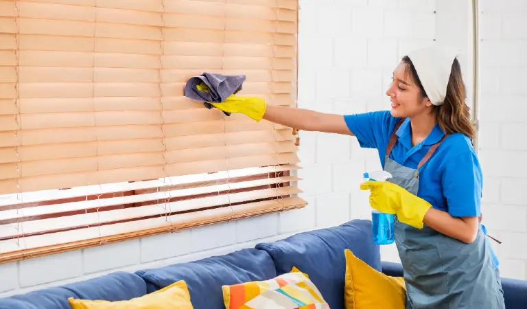 Smiling professional cleaner in blue uniform wipes window blinds in a modern living room