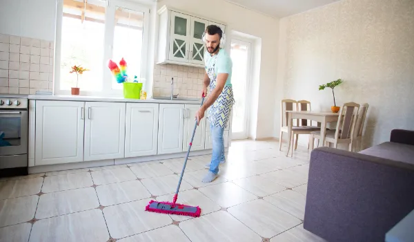 Happy young beard man is cleaning the floor in the domestic kitchen and having fun